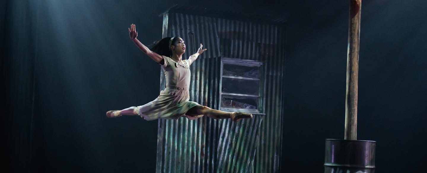 Dancer wearing a light dress leaps gracefully high above the stage, the set of a ramshackle building behind her