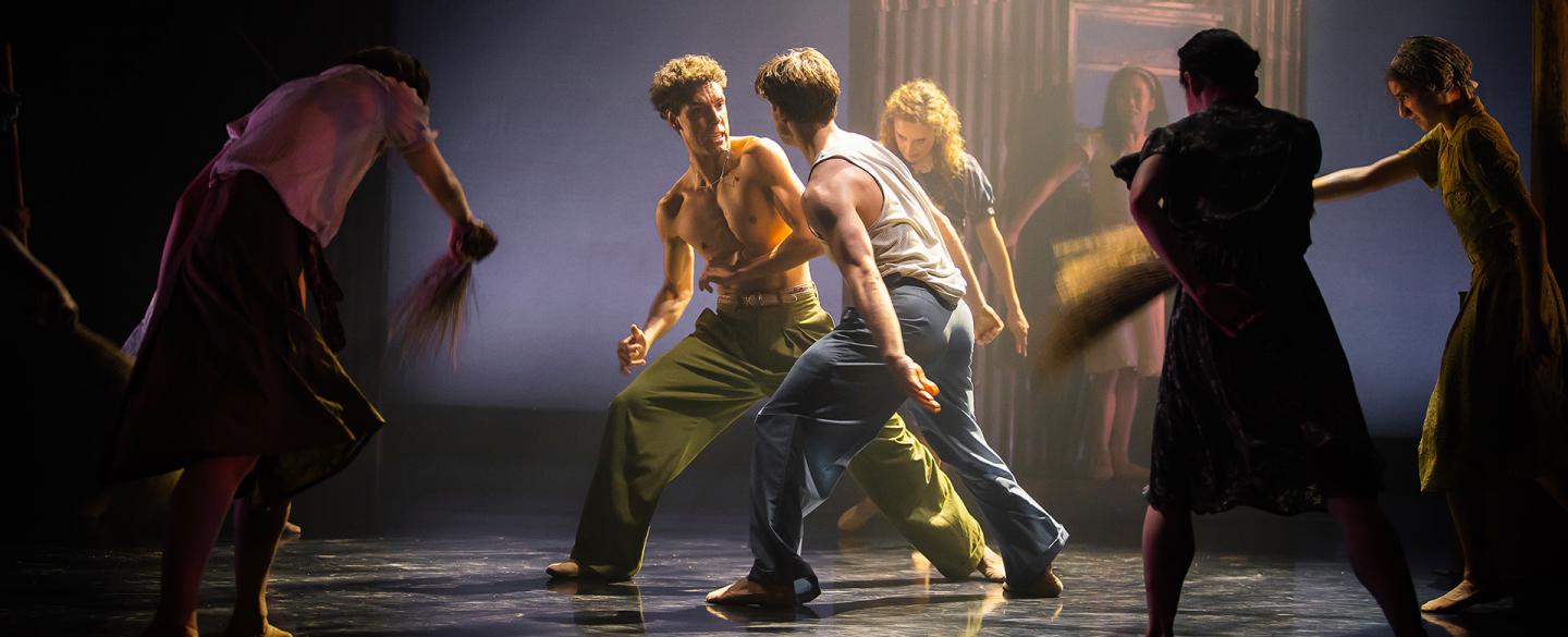 Two dancers, one bear chested in green trousers, the other in blue trousers with a white vest, fighting, one turned slightly and leaning back with an angry expression about to punch the other