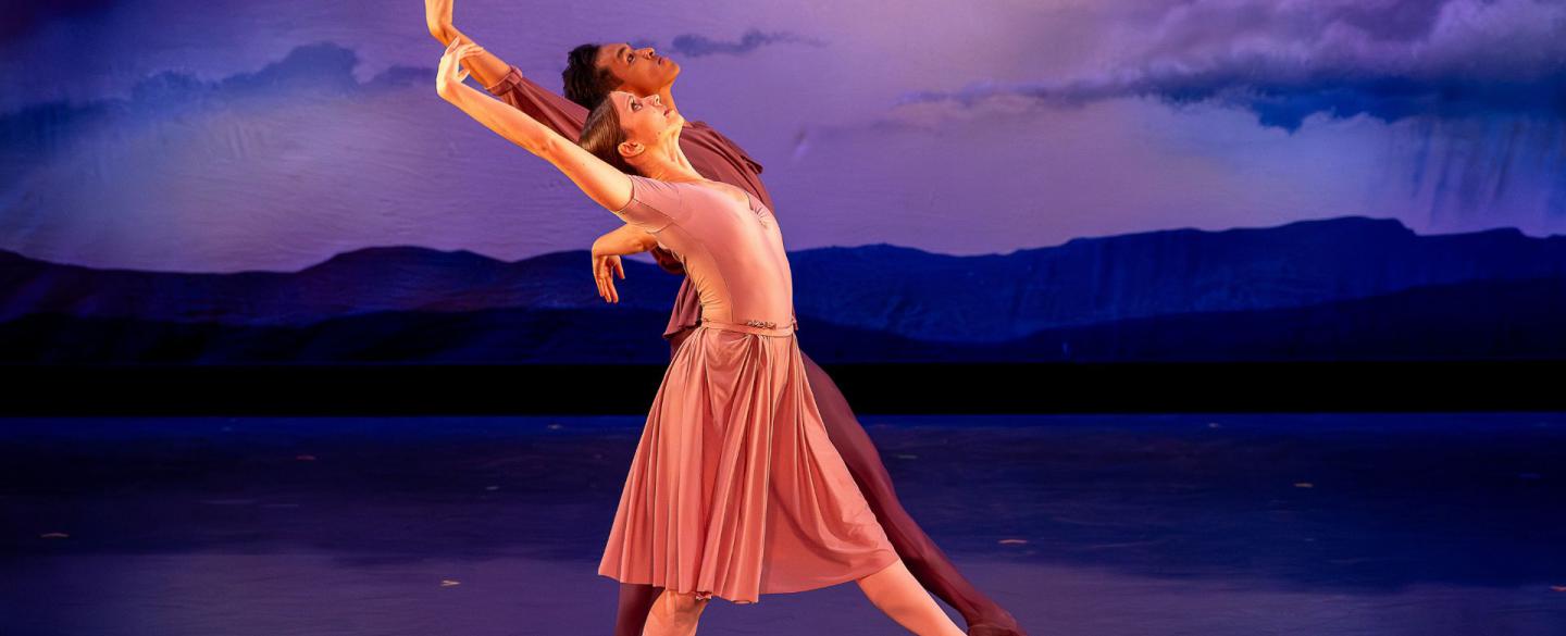 Two dancers against a cloudy background move to the right while leaning backwards and lifting their arm above their heads.