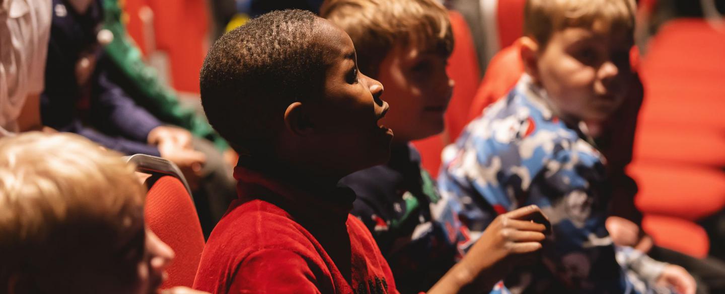 Children in Christmas jumpers sit in an auditorium. One boy opens his mouth to speak to someone out of frame.