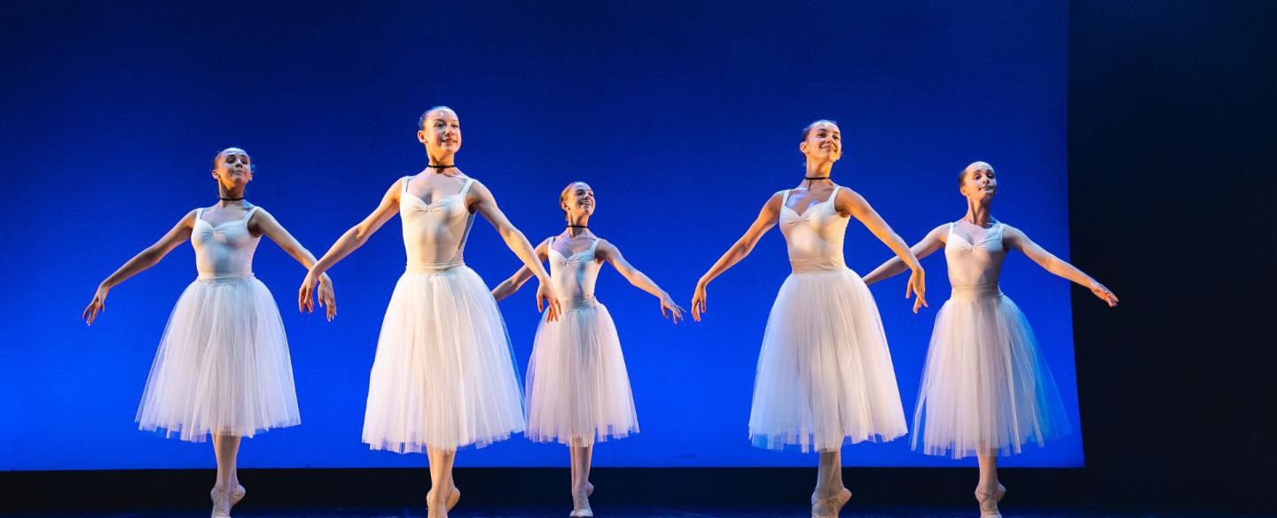 Students in white perform on a stage, standing on their toes with their arms at their side.