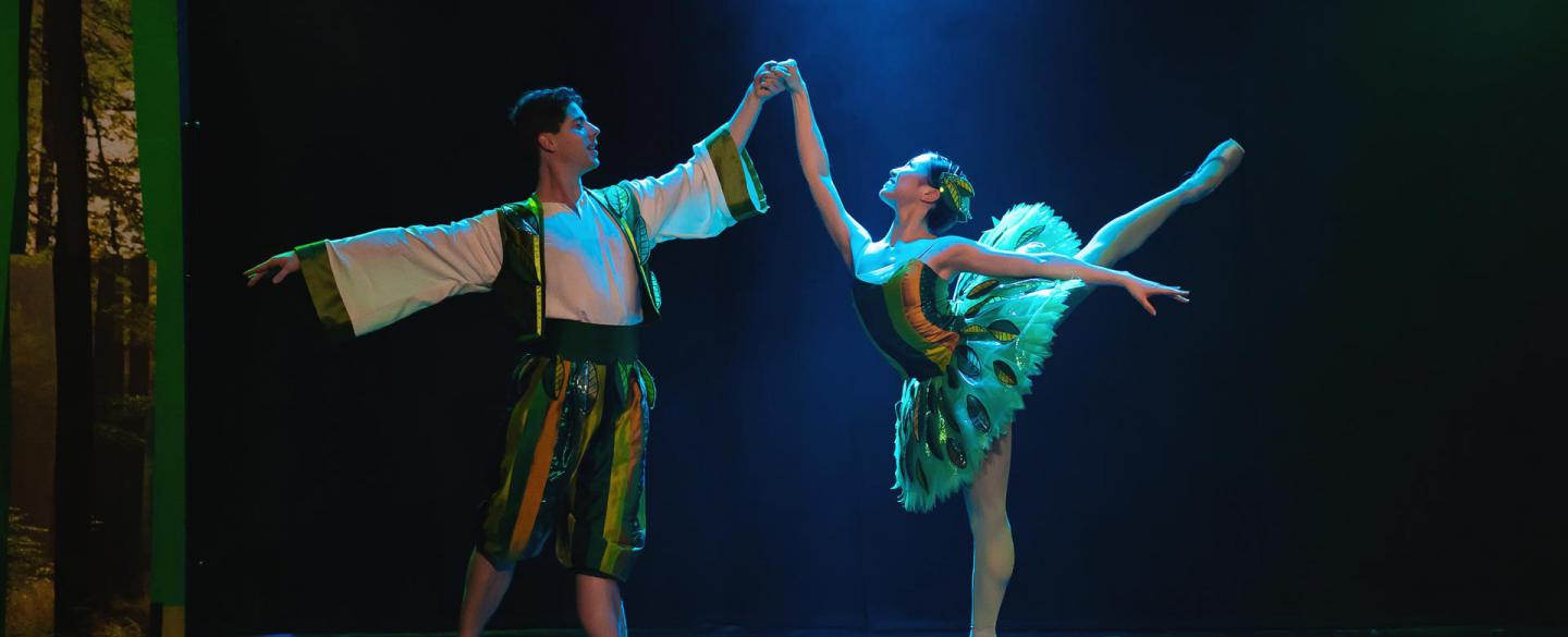 Two dancers stood in a traditional ballet pose. One dancer is on pointe with leg raised high