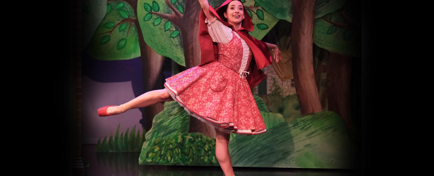 Dancer in red with a red hood and red pointe shoes stands arabesque smiling as she walks through a forest