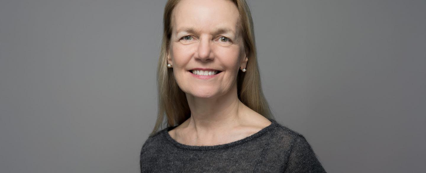 A headshot of a white women with blonde hair wearing a black top smiling to camera against a grey background.