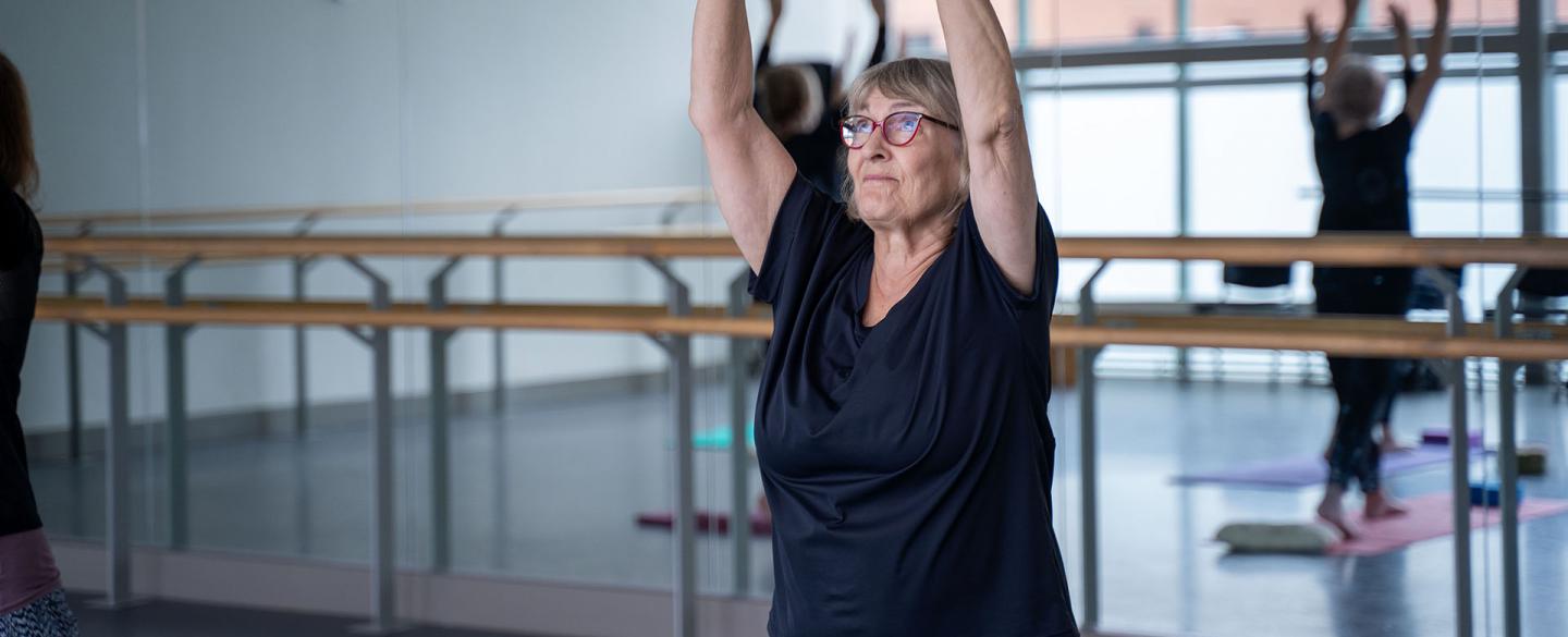 Student in glasses and dark T-shirt, arms raise above her head, participating in a class for people over 55