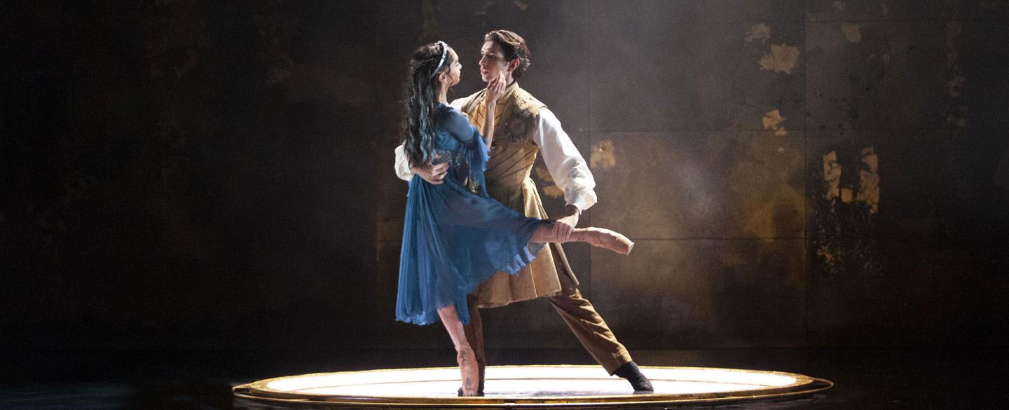 In an illuminated circle two people dance, a woman en pointe in a blue dress and a man who his holding her lovingly