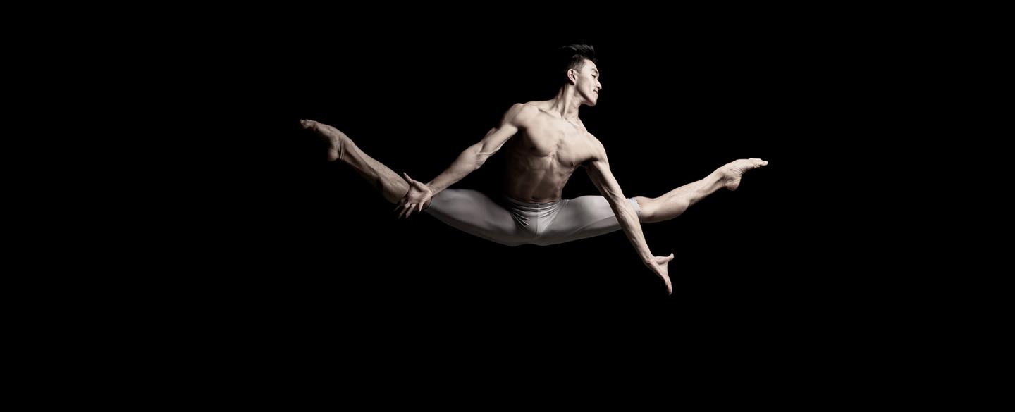 Male dancer, only wearing white leggings, mid jump, legs split, arms reaching down