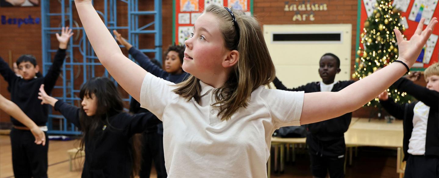 Young girl with straight shoulder-length hair stands in a school hall with her class-mates, arms wide, hands raise and smiling