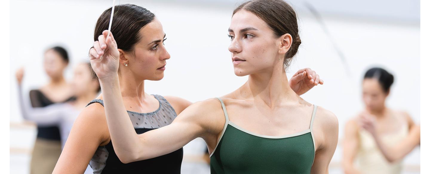 Dancer with feather in hand looks over her shoulder at another placing a hand on her shoulder attentively