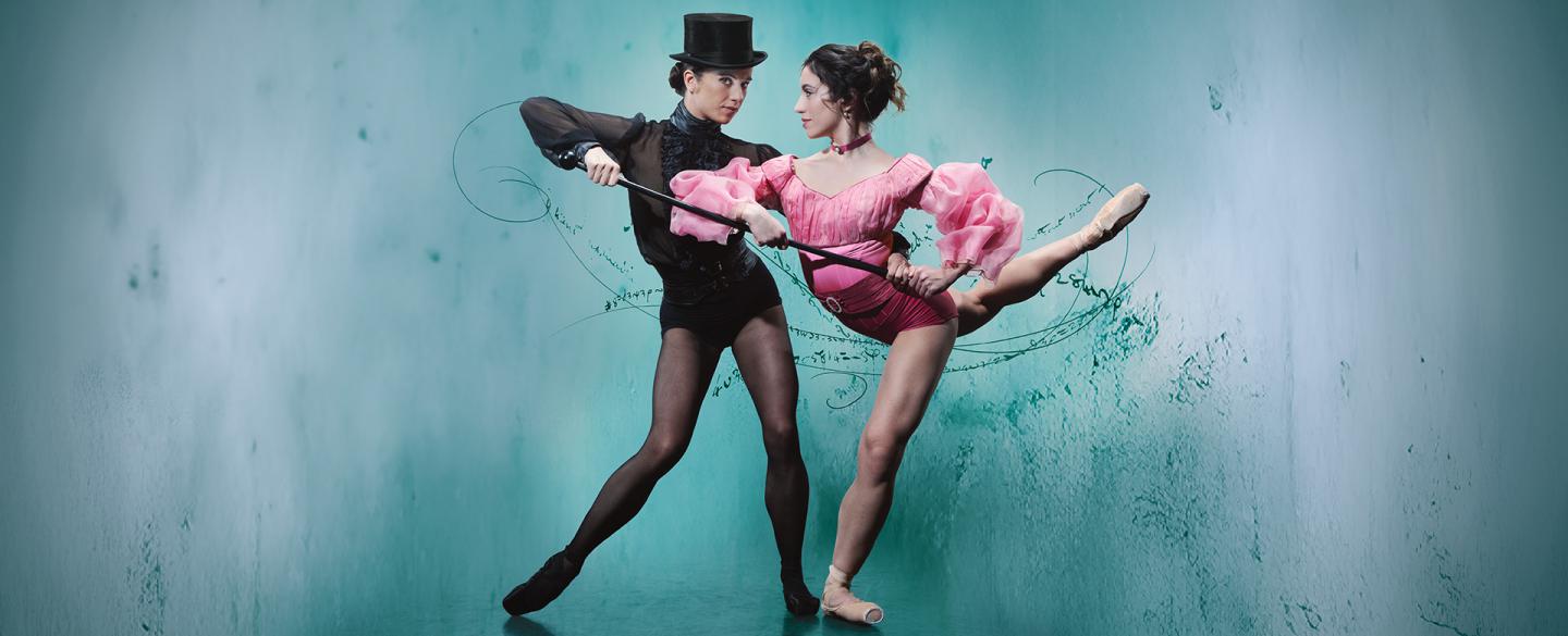 Two women, one in black with black pointe shoes and hat supporting another woman with a walking stick, her wearing pink