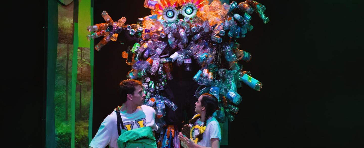 A dancer in a costume covered in plastic bottles sneaking up on two dancers sat at the front of the stage.