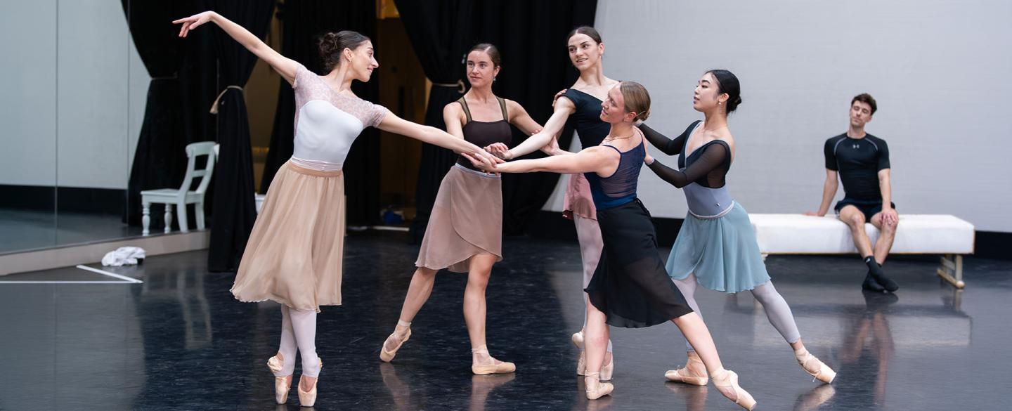 Five dancers in the rehearsal room in a circle all touching hands in the centre, one en pointe