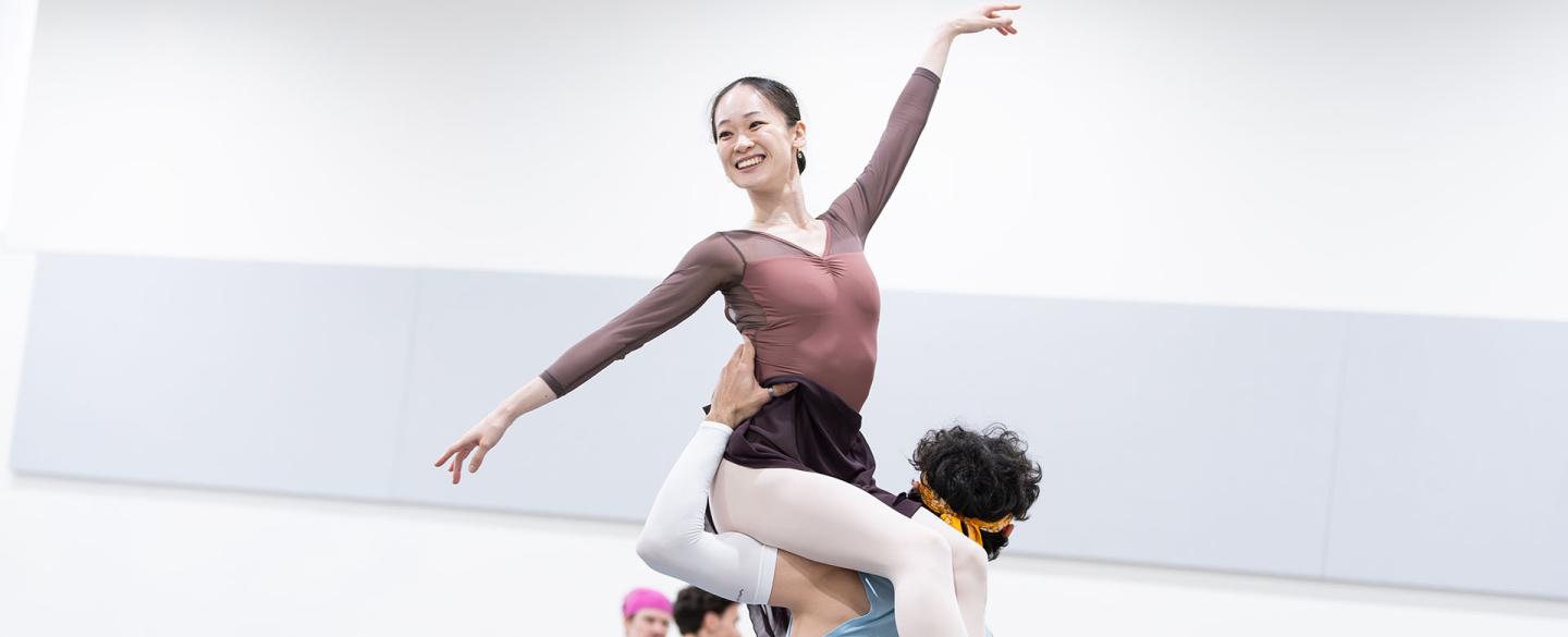 Dancer smiling as she is lifted, sat on the left shoulder of another dancer, one arm angled up, the other angled low