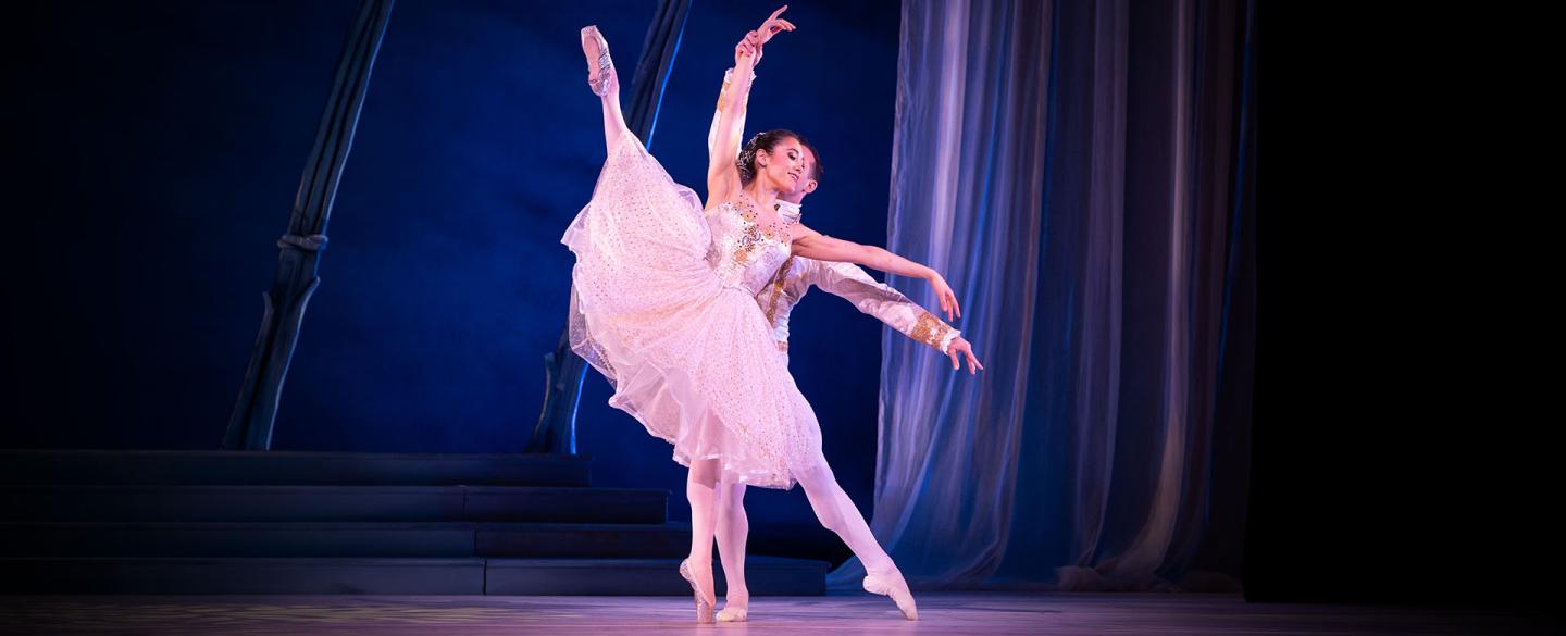 Woman in a shite flowing skirt and satin top stands on one foot en pointe and the other leg is exteded into the air, another dancer stands behind her supporting her balance.