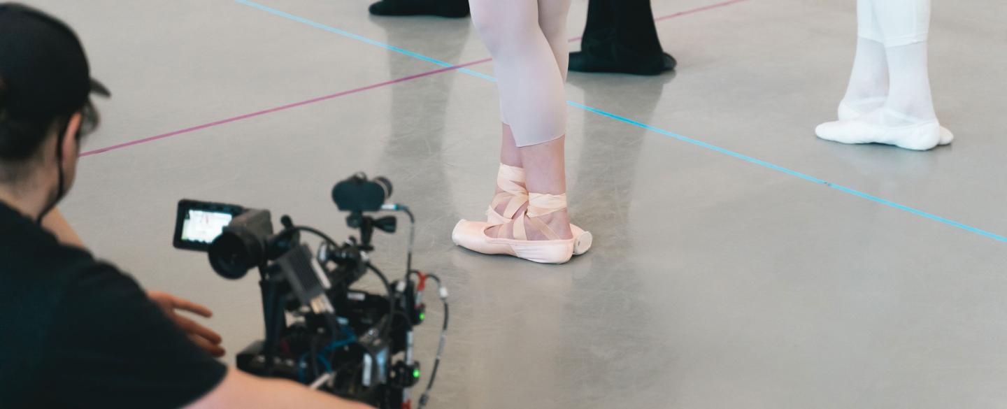 A camera operator in black focusing on a dancer in pointe shoes with legs crossed in a fifth position