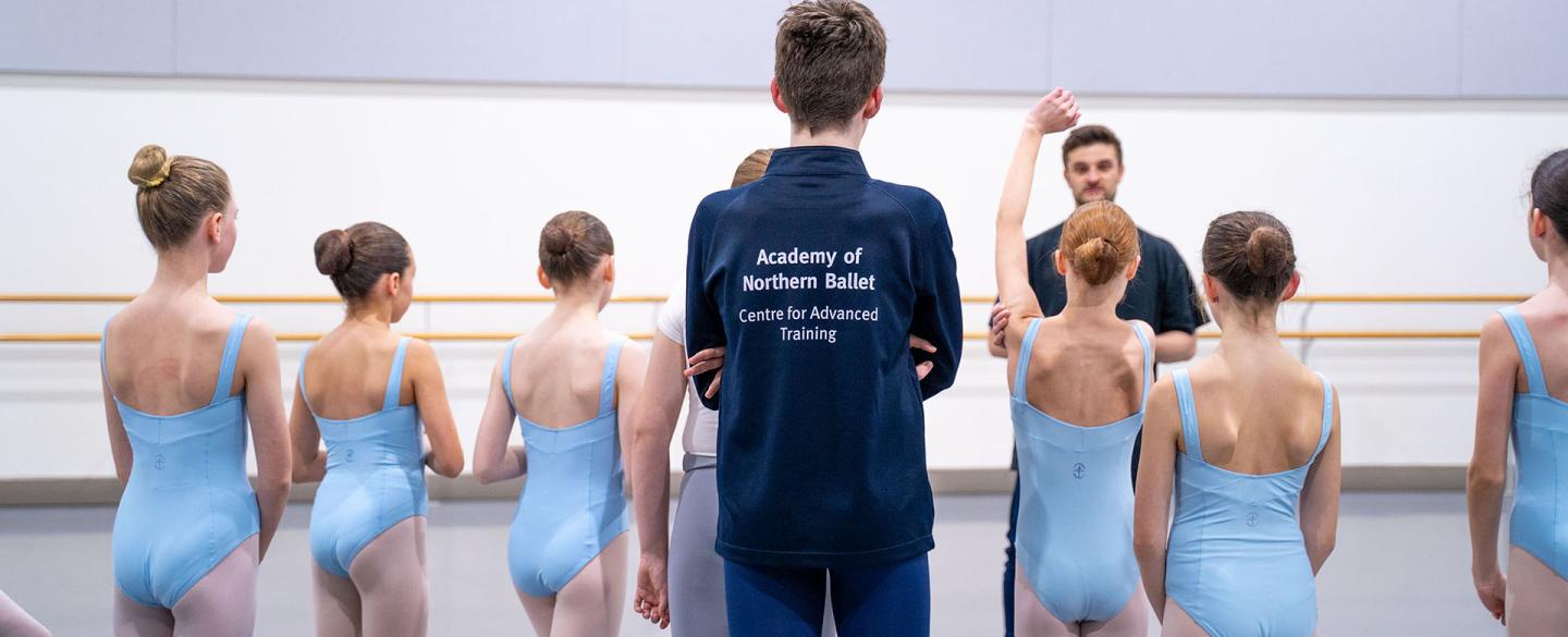Group of Centre For Advanced Training students with their backs to the camera, dressed in blue leotards standing in front of their teacher, one with a raised hand to answer a question