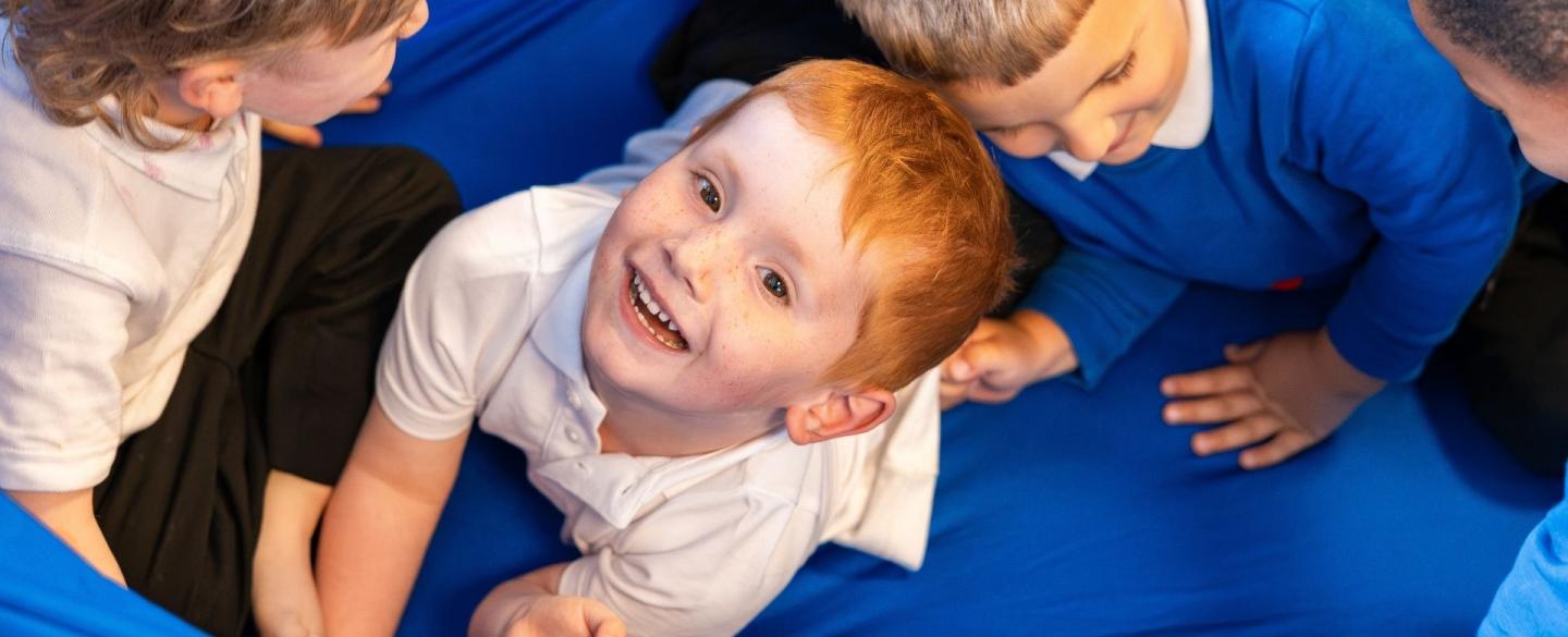 Three young school boys play on top of a bright blue fabric. The boy in the middle looks up at the camera with a smile.