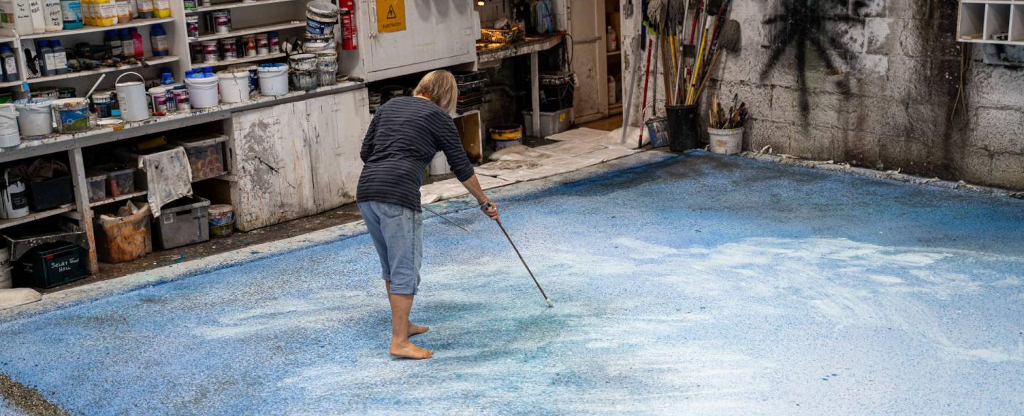 In a workshop, a person wearing a black top and jeans stands on a large canvas painting with a long paintbrush.