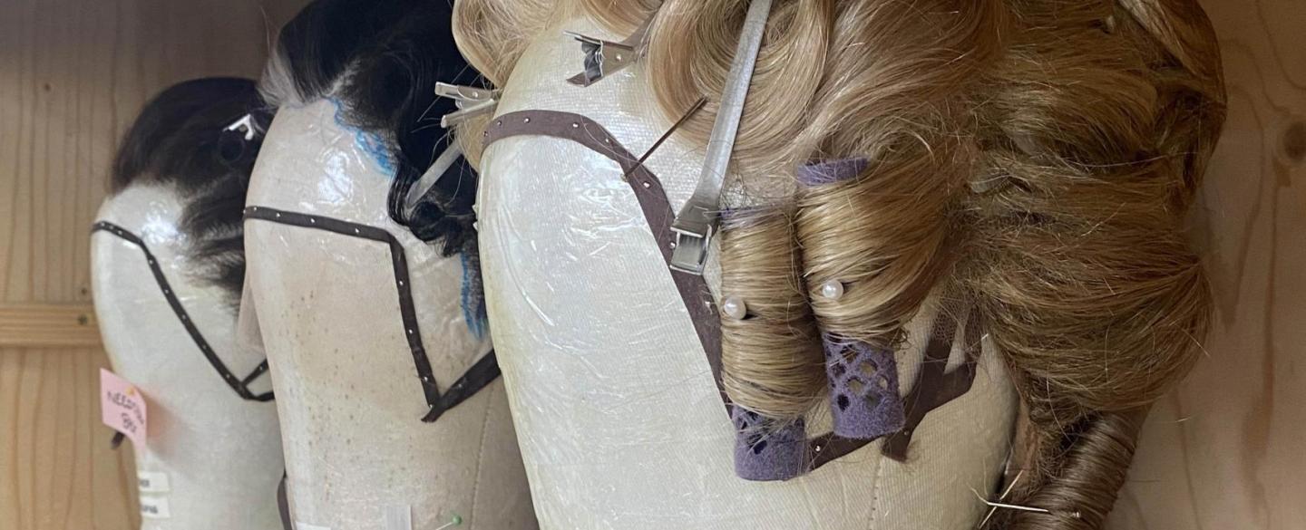 A row of 3 wigs are on display after being styled. There is one blonde and two brunette. They have rollers, pins and clips in them.