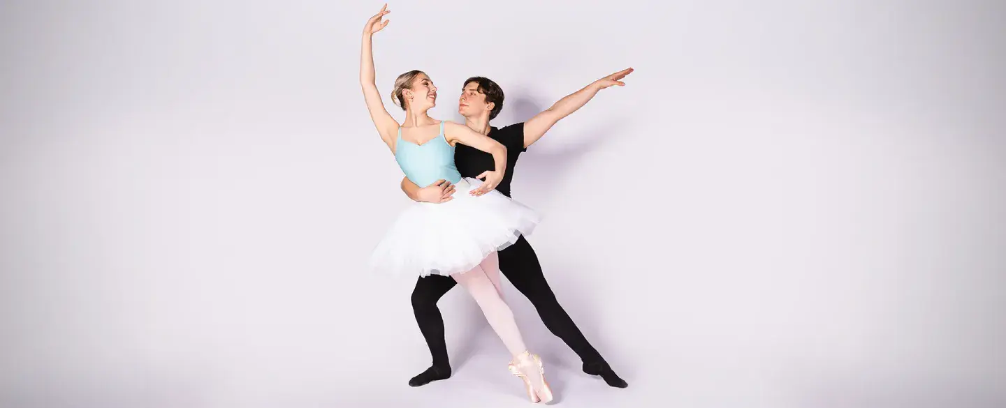 Two student dancers against a pale pink backdrop, one in a blue leotard and tutu on pointe and leaning supported by the other all in black with an arm around her waist