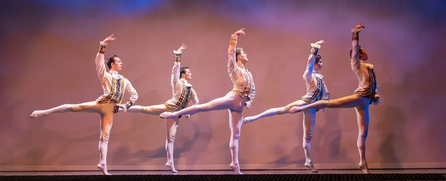 Five male dancers in white and bodies in sync, standing straight up on one foot, one hand on hip, the other arm high in the air