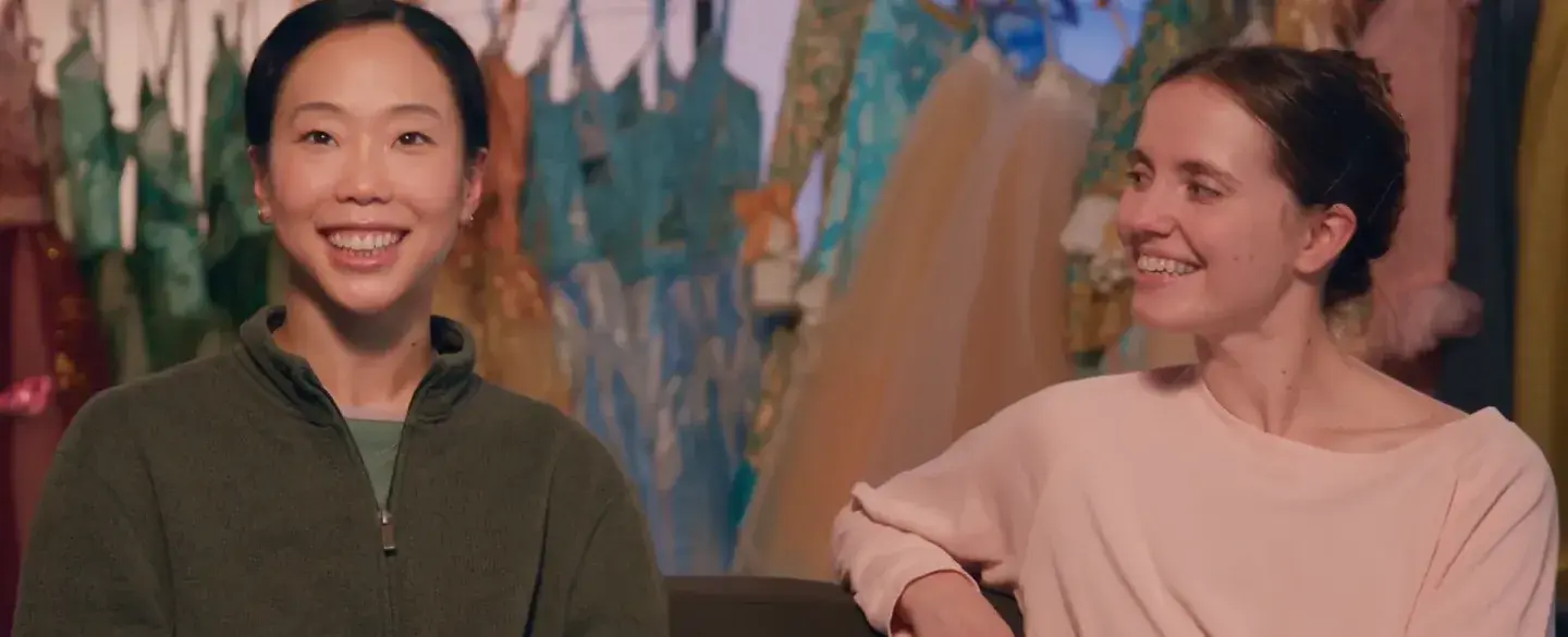 Two people sat on a sofa smiling with costumes hung on a rail in the background