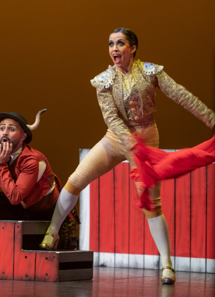 Two dancers on a stage, surrounded by red pieces of set including a small stair case. One dancer is nelt down behind the stair case wearing a red jacket and bull ears on their hat. They look shocked. The other dancer is stood up with their right foot on the staircase, wearing a gold outfit, looking shocked and they have pulled away a red cape.