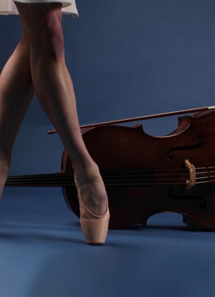 Photo of the bare lower legs wearing pointe shoes in front of a viola places on its side all against a blue background