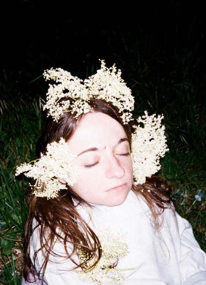 PErson on grass in the dark with her eyes closed, lit brightly with white flowers in her hair and a wearing white shirt