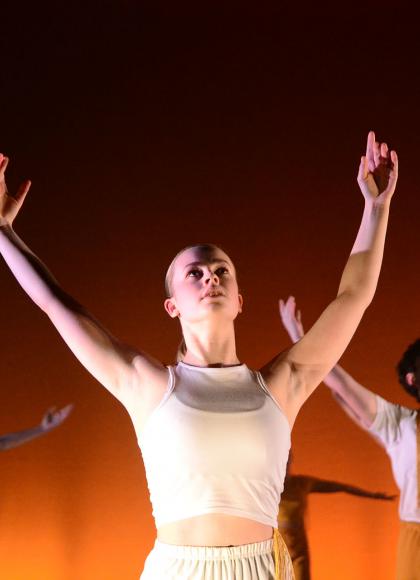 Young female dancer in a white top against a lit orange background looks into the air and raises her hands above her head