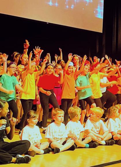 Group of over 50 children wearing different colours all waving out from a stage happily