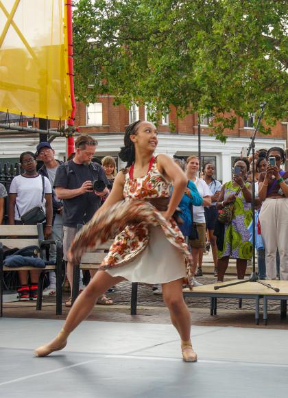 Two black dancers, one in a bright flower dress, the other in dark trousers and T-shirt, dance on stage while members of the public look on