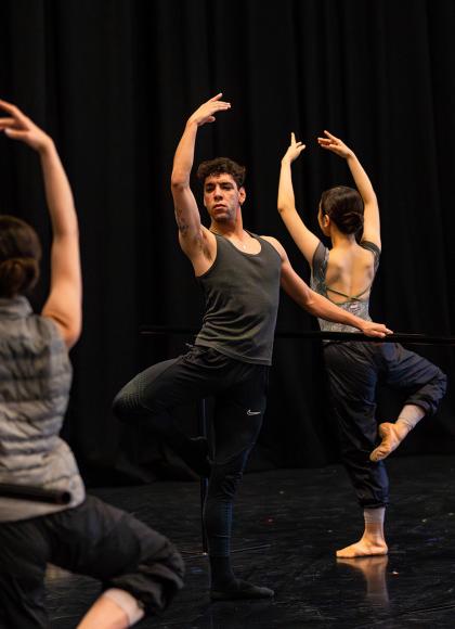 Dancer in a dark vest, one hand hovering above a barre, the over raised above his head, standing on one foot with the other leg raised at the knee