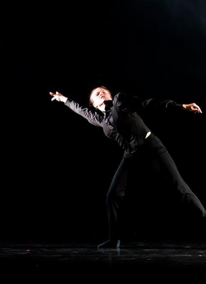 Dancer in a dark top and trousers, against a darkened stage, stands with legs apart, leaning to their right with an arm outstretched