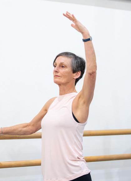 Standing at the barre with one hand holding on, a student of the Over 55s class wearing a pink top raises her other arm high while focusing in front of her