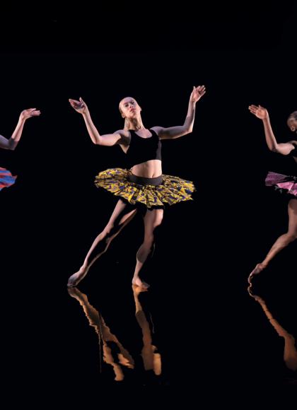 Against a back background and shiny black floor, three dancers dressed in colourful tutus pose with their arms raised, bent at the elbow