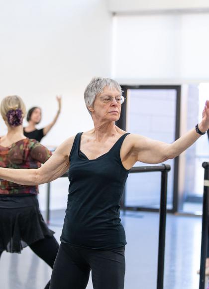 Class of dancers at the barre, one hand holding on for balance, the other arm held out, palm up forearm raised, knees slightly bent