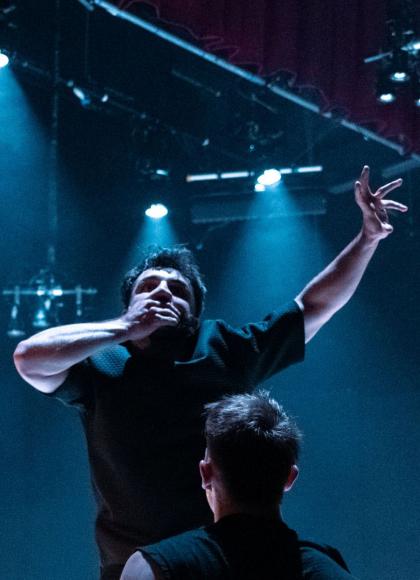 Two dancers stand on a dark stage lit by intense blue spotlights. One dancer stands with his back to the camera, while the other faces forward, covering his mouth with one hand and raising one arm in an expressive gesture.
