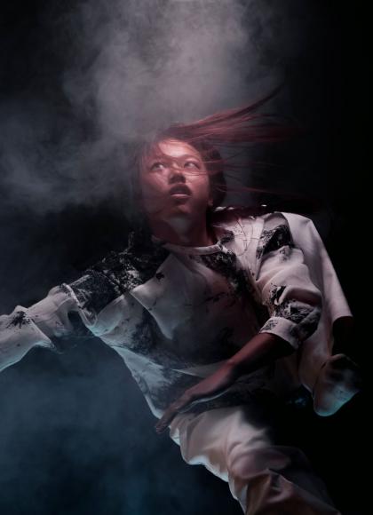 On a black background, a dancer is captured in mid-motion, with their body suspended in the air and surrounded by a swirl of smoke. 
