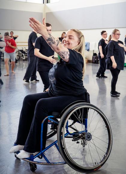 Woman in wheelchair smiling and raising her arm expressively. Young people in the background moving in unison