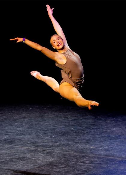 Woman on a stage performing a grand jete wearing a brown top and shorts, slicked back hair and an open smile