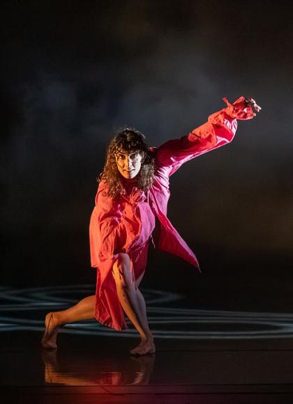 On a dark stage, a woman in pink is pictured. She has her right leg planted on the floor with her left leg crossing behind it, making her lunge. She is looking forwards with her left arm up in the air and her right close to her chest.