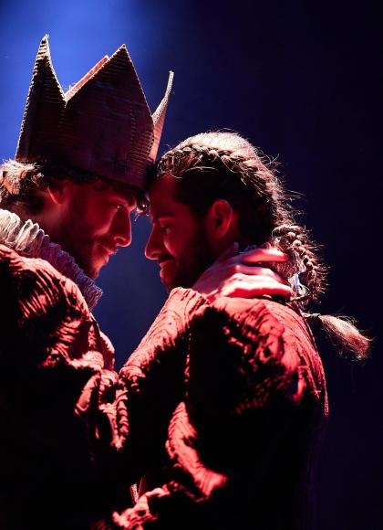 On a dark background, two male dancers are facing each other in an embrace. Their foreheads are touching and they are looking into each other's eyes. One of the men is wearing a large crown on his head.