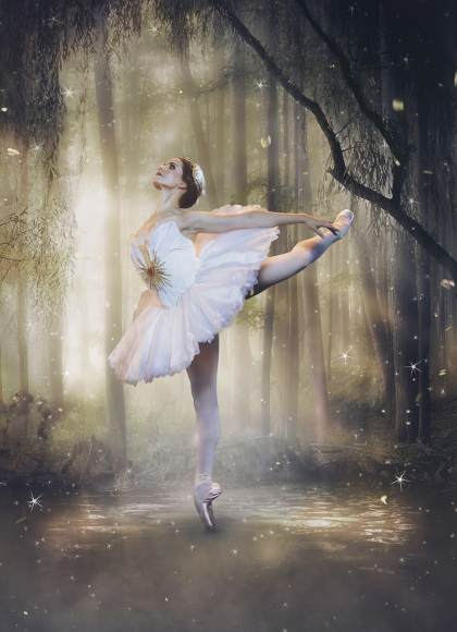 In the middle of a dark forest with soft sunlight, a dancer in a white feathery tutu poses in an attitude, with her back arched and her arms reaching to the back.