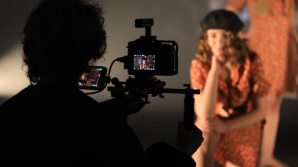 Behind the scenes at the filming of a dance film with students. We see the silhouette of the camera operator and the back of the camera, in the background we see a student looking down the camera, out of focus,