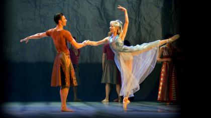 Two dancers on stage, one supports the other as she stands en pointe.