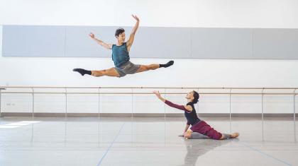 A male dancer leaps through the air in a full jete while a female dancer reaches up to him while sat on the floor