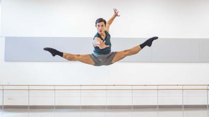 A male dancer jump with both legs extended in a mid-air split while reaching for the camara