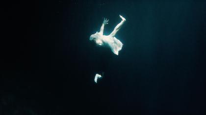 A man in a white shirt and dark trousers shapes his body like a swan whilst underwater