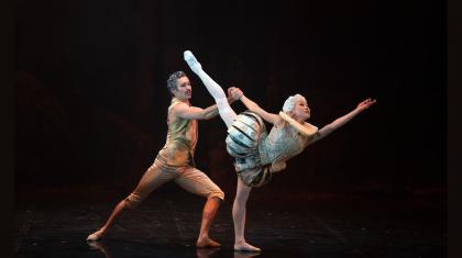 A female dancer in light 18th century costume reaches into the distance with her leg aloft, supported from behind by a male dancer in gold costume.